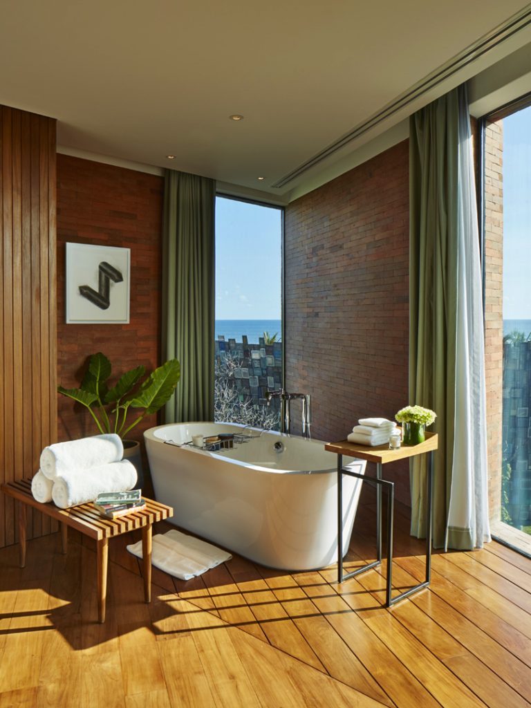 The bath area in the Potato Head Suites at Katamama Hotel in Seminyak, Bali features a bathtub and a bench with towels.
