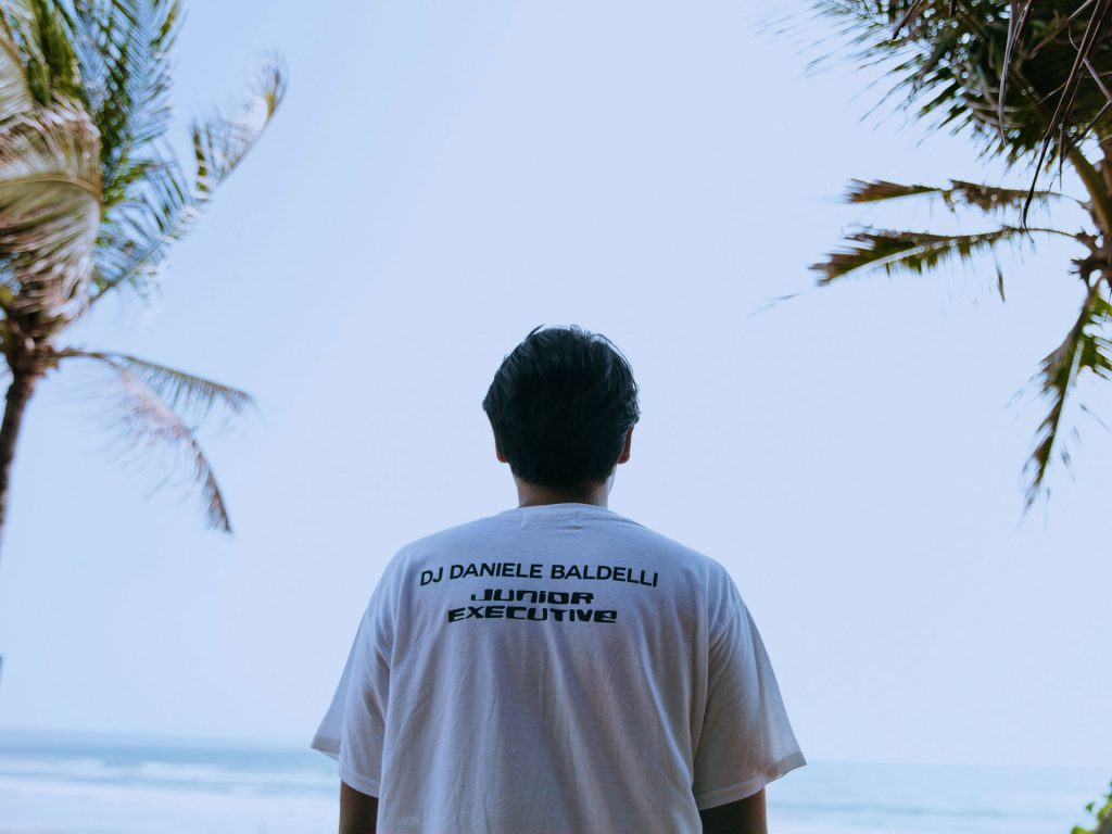 A man wearing DJ Daniele Baldelli x Junior Executive white t-shirt at the beachfront of Desa Potato Head, Seminyak, Bali