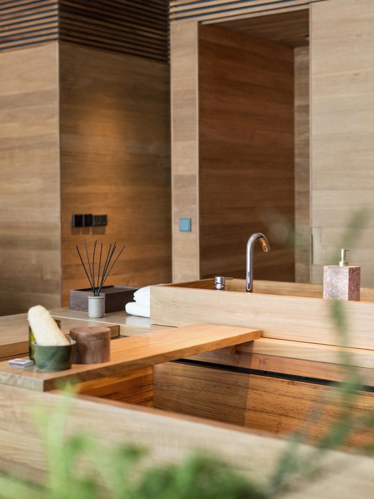 The wooden bathroom of the Oceanfront Studio at Potato Head Studios features a bath tub and a sink with a reed diffuser.